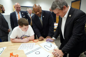 An ATI-UMW ninth-grader demonstrates a STEM activity for UMW Board of Visitors Rector Lee Murray (orange tie), BOV member Terris Todd (center) and Del. Phillip Scott, along with other tour-takers before the recent dedication ceremony. Photo by Suzanne Carr Rossi.